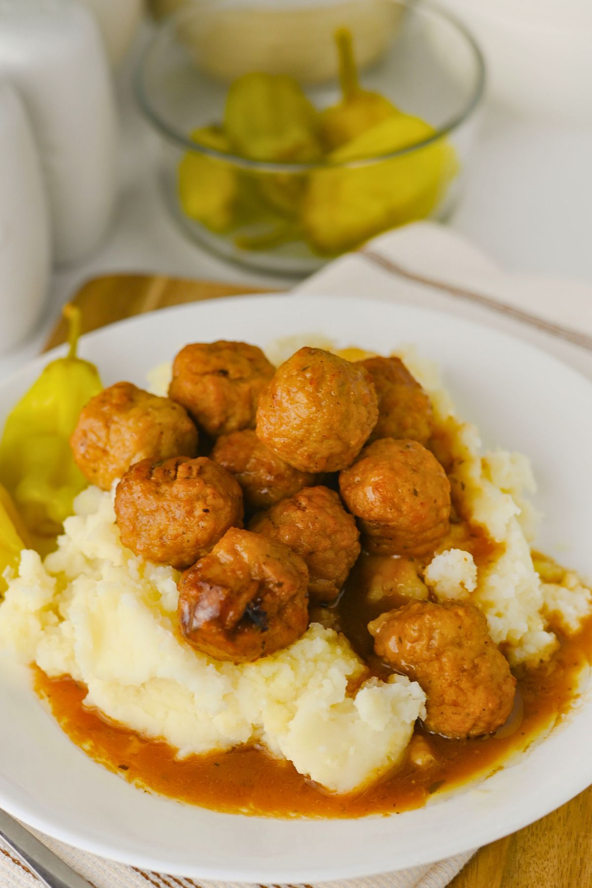 A plate of mashed potatoes topped with meatballs in brown gravy, served with a yellow pepper on the side.