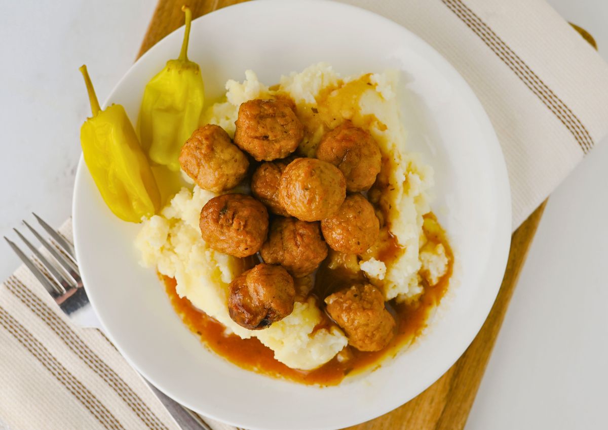 A white plate with mashed potatoes topped with meatballs in brown gravy, served with two yellow peppers on the side.