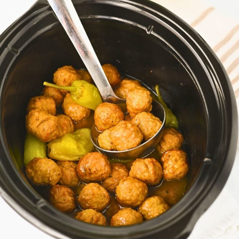 A slow cooker filled with meatballs, pepperoncini peppers, and sauce, with a metal ladle scooping some meatballs.