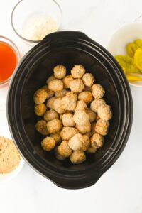 A slow cooker filled with frozen meatballs, surrounded by bowls containing breadcrumbs, red sauce, banana peppers, and a powdered ingredient.