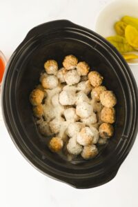 Frozen meatballs coated with grated cheese and seasonings inside a black slow cooker, with a bowl of yellow pepper slices visible nearby.