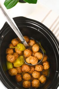 A slow cooker filled with meatballs, pepperoncini peppers, and broth, with a metal ladle resting inside.