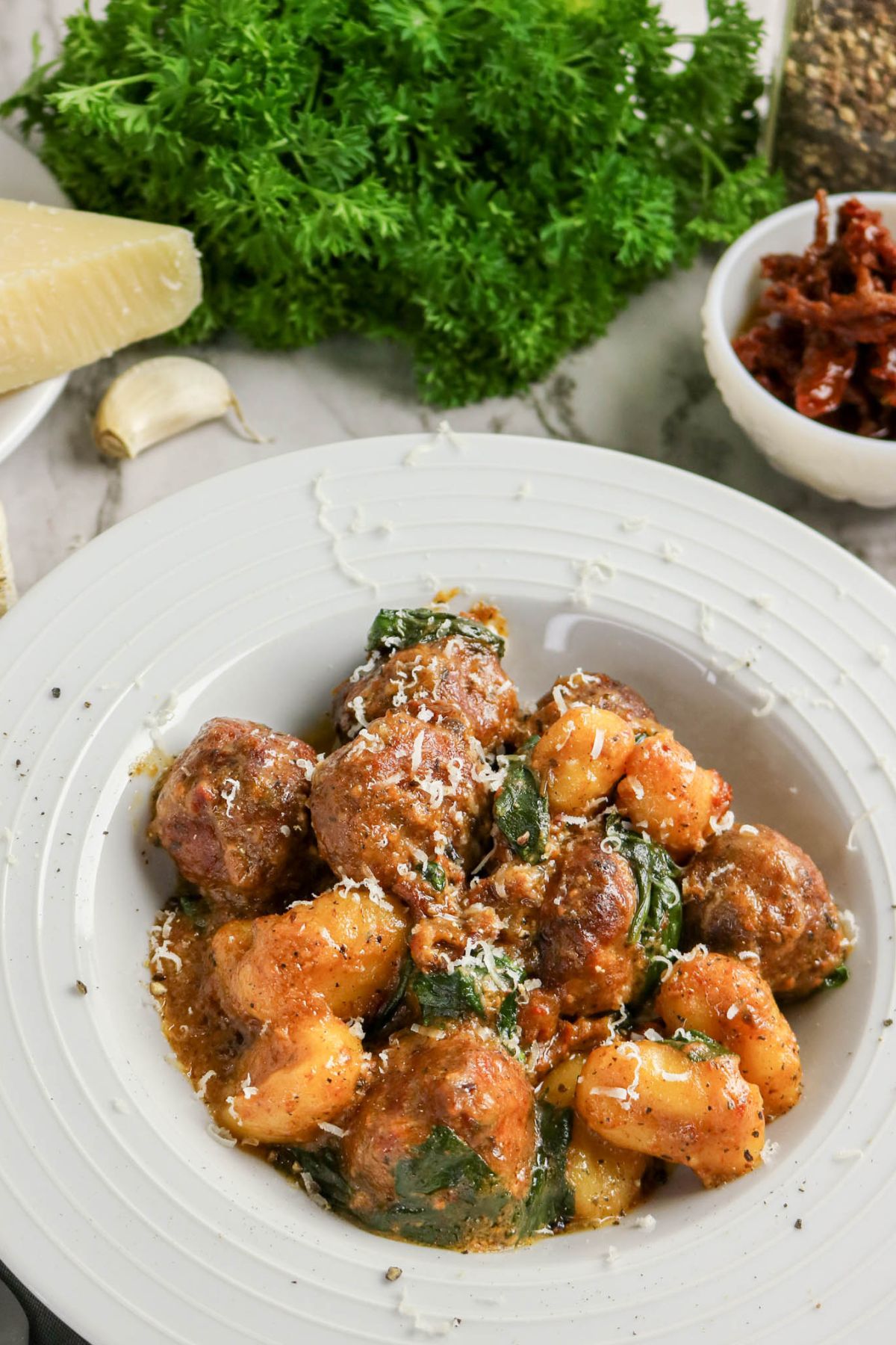 A white plate of meatballs, gnocchi, and spinach in tomato sauce, garnished with grated cheese, with parsley, parmesan, garlic, and sundried tomatoes in the background.
