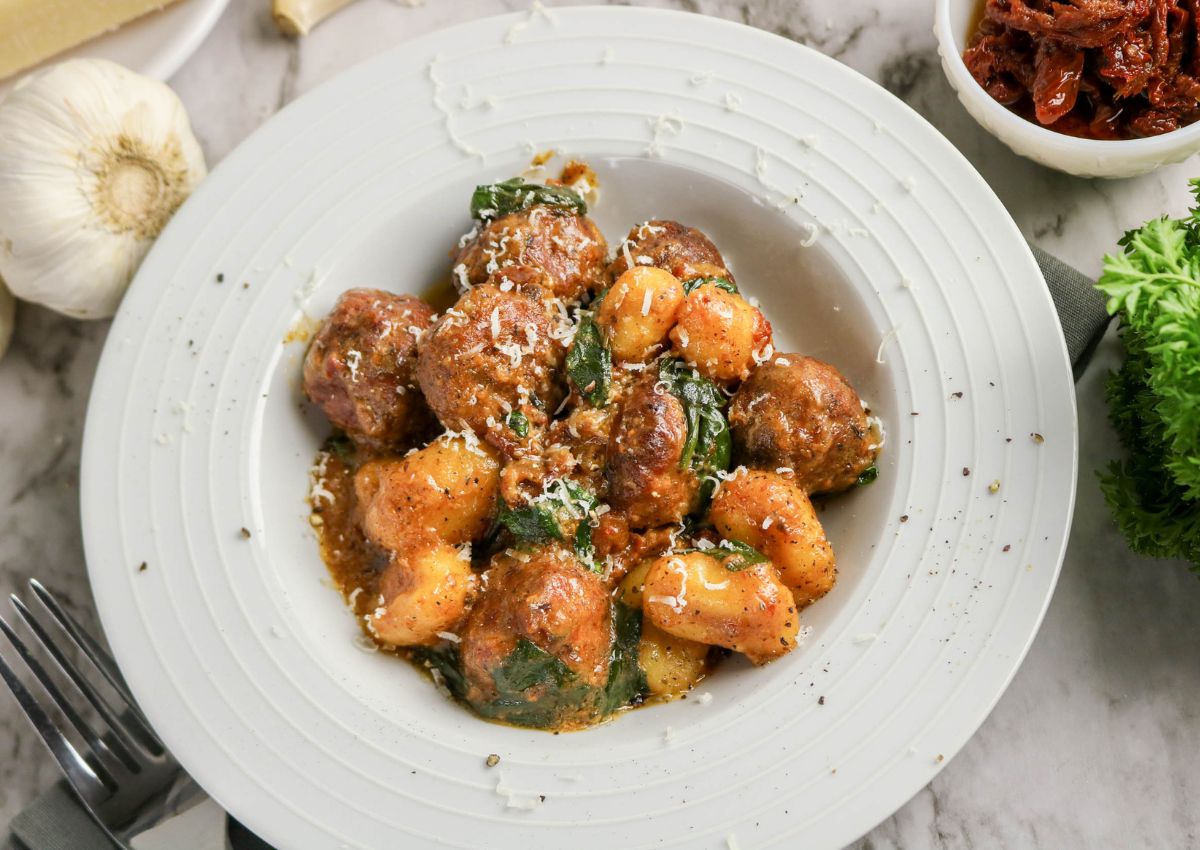 A white plate of gnocchi and meatballs in tomato sauce, topped with grated cheese and herbs, placed on a marble surface with garlic, parsley, and sun-dried tomatoes nearby.