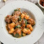 A white plate of gnocchi and meatballs in a tomato-based sauce, topped with grated cheese and garnished with spinach.