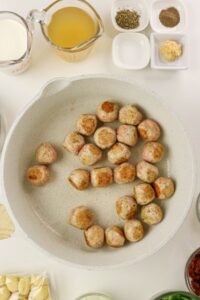 Browned meatballs cooking in a white skillet, surrounded by ingredients in small bowls, including broth, cream, garlic, spices, and gnocchi.