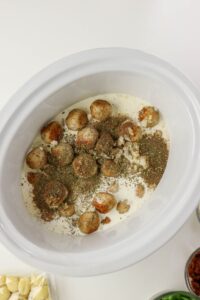 A slow cooker filled with cream, meatballs, herbs, and seasonings, shown from above before cooking. A package of gnocchi and bowls of ingredients are nearby.