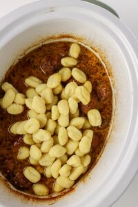 Uncooked gnocchi sits on top of cooked meatballs and sauce inside a white slow cooker.