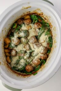 A slow cooker filled with meatballs, beans, spinach, and topped with grated parmesan cheese.