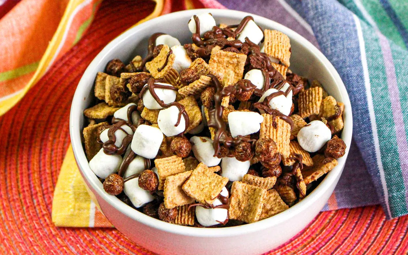 A bowl filled with cereal squares, chocolate pieces, cocoa puffs, marshmallows, and drizzled with chocolate, set on a colorful woven placemat with napkins nearby.