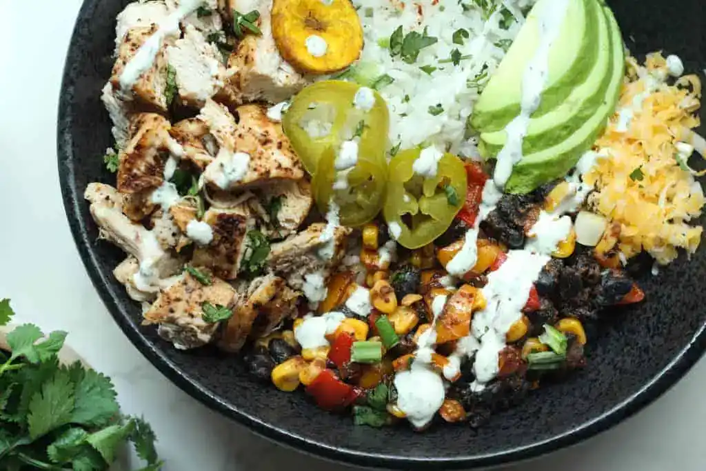 A black bowl filled with grilled chicken, white rice, avocado slices, shredded cheese, black beans with corn and peppers, plantain, jalapeรฑo slices, and a drizzle of white sauce.