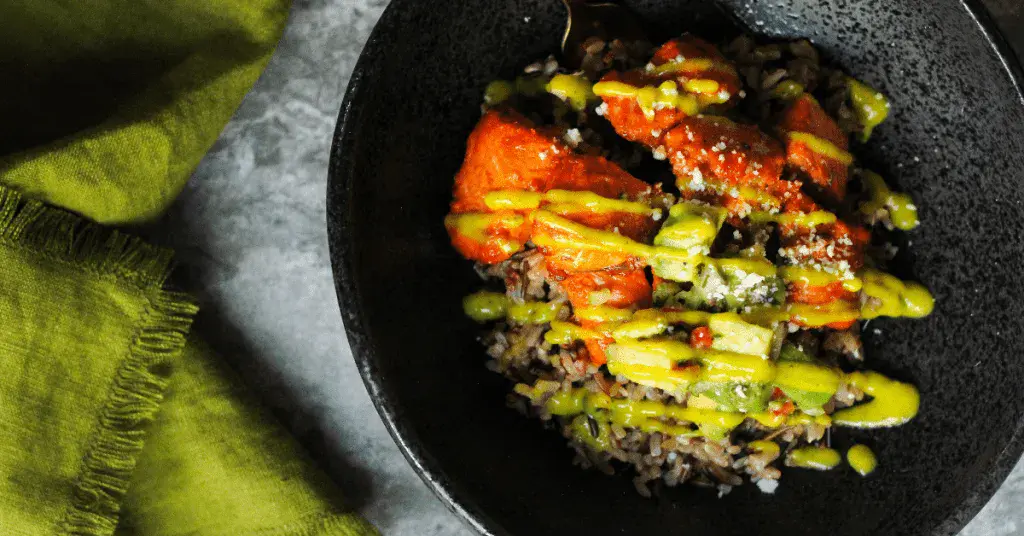 A black bowl containing rice topped with pieces of orange-colored food and drizzled with green and yellow sauce, next to a green cloth napkin.