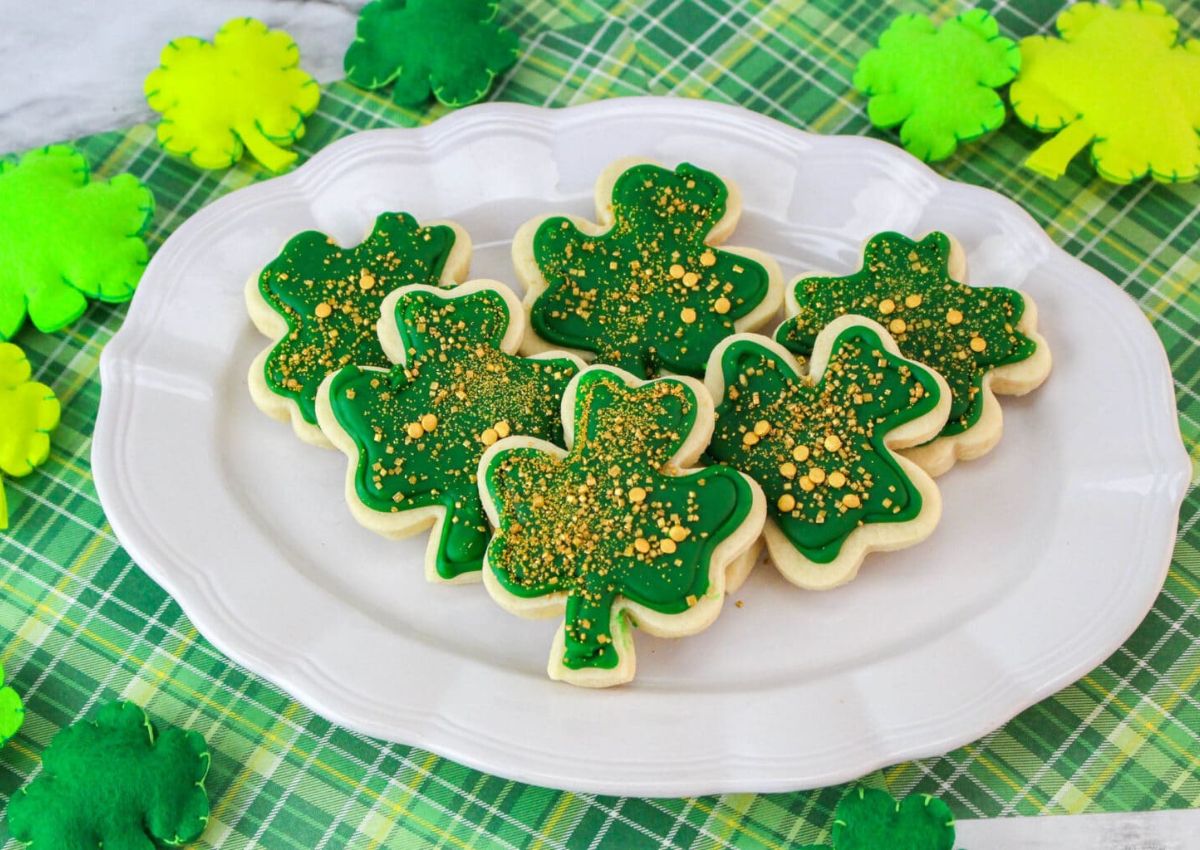 Shamrock-shaped cookies decorated with green icing and gold sprinkles are arranged on a white plate, with felt shamrock decorations and a green plaid cloth underneath.