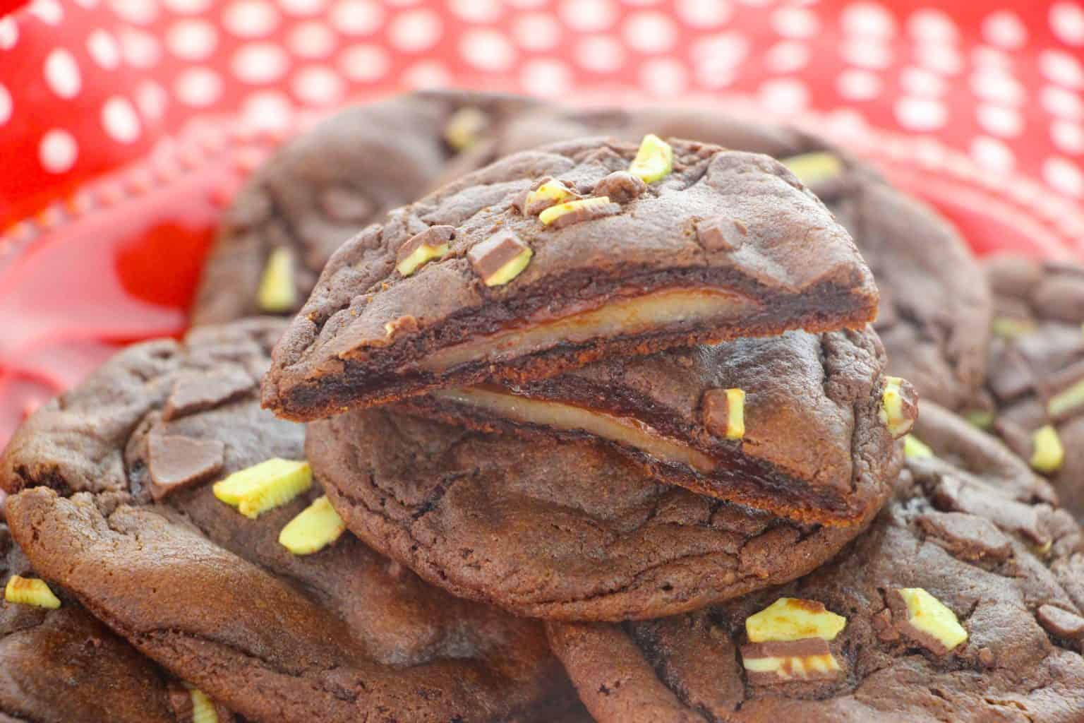 A close-up of chocolate cookies with a creamy center, topped with small pieces of yellow and brown candy, stacked on a red plate with a white polka-dot background.