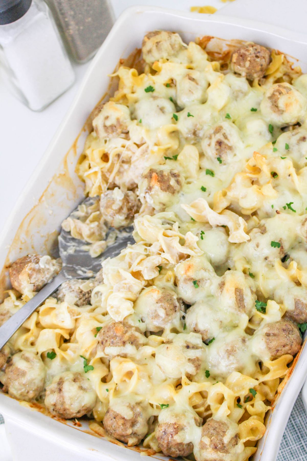 A casserole dish filled with baked noodles, meatballs, and melted cheese, with a serving spoon and fork partially inserted.