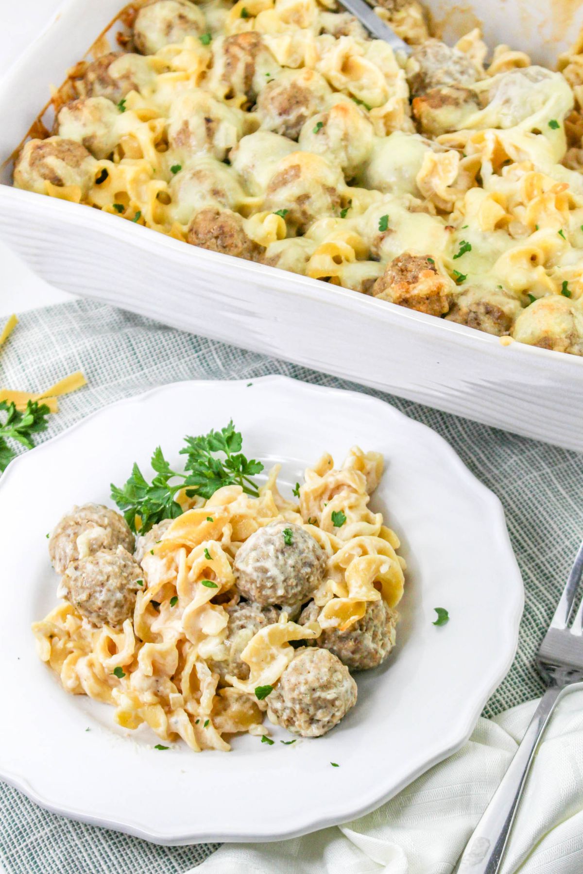A white plate with egg noodles and meatballs in a creamy sauce, garnished with parsley, beside a baking dish of the same noodle and meatball casserole topped with melted cheese.