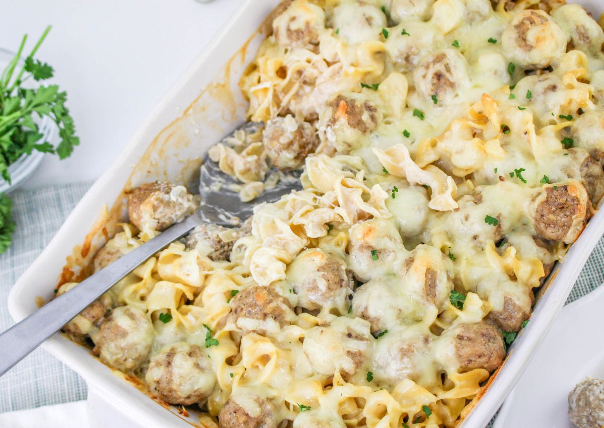 A casserole dish filled with baked noodles, meatballs, and melted cheese, with a serving spatula and some chopped parsley on top.