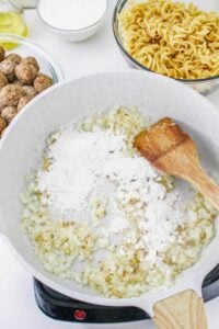 Chopped onions and flour being sautéed in a white skillet with a wooden spatula; bowls of uncooked noodles, meatballs, and a white sauce are in the background.