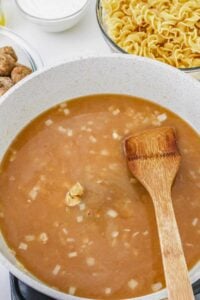 A skillet with brown gravy and chopped onions being stirred by a wooden spatula; uncooked egg noodles, meatballs, and a bowl of sour cream are nearby.