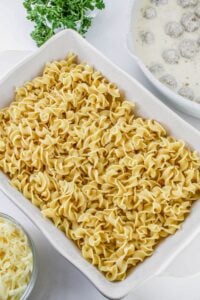 A white baking dish filled with uncooked egg noodles, with shredded cheese, a parsley bunch, and a bowl of meatballs in cream sauce nearby.