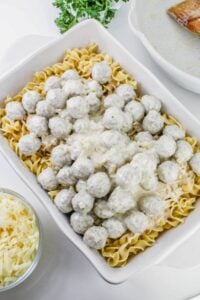A white baking dish filled with cooked egg noodles topped with meatballs and creamy white sauce. A bowl of shredded cheese and a sprig of parsley are nearby.