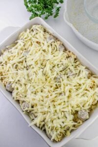 Rectangular baking dish filled with pasta, sausage pieces, and topped with shredded cheese; mixing bowl and parsley in the background.