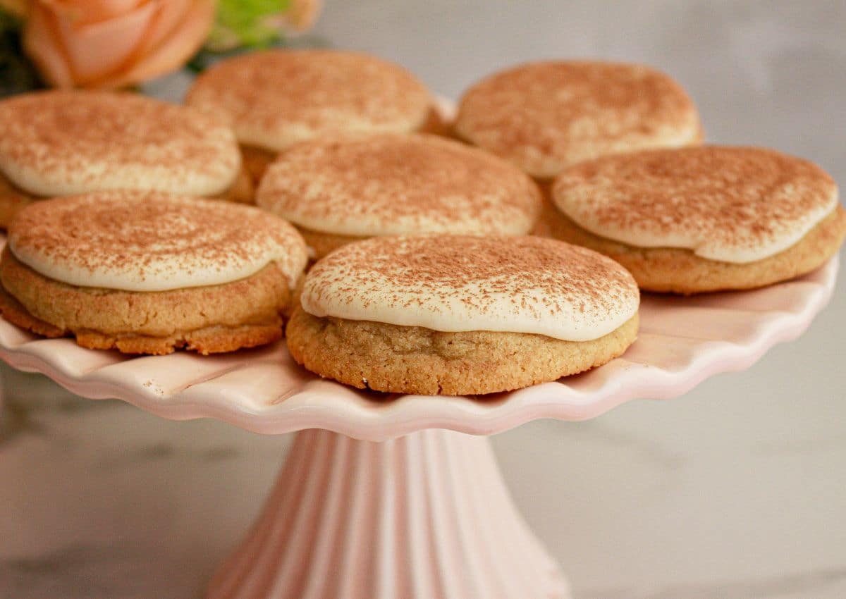 Seven cookies topped with frosting and a dusting of cocoa powder are arranged on a light pink cake stand.