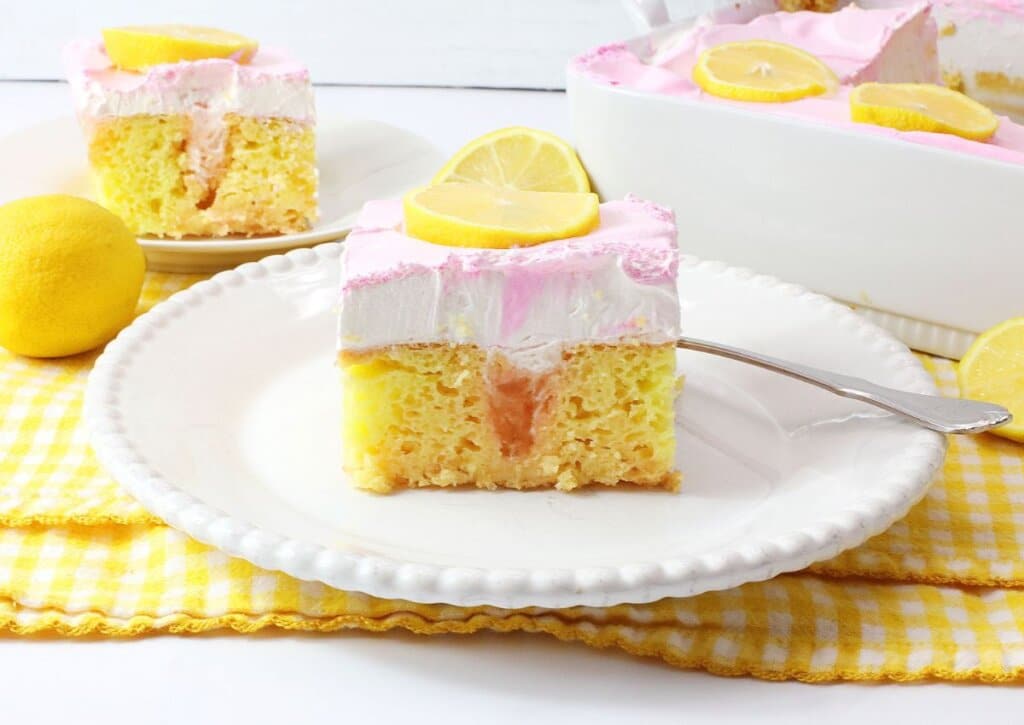 A slice of yellow lemon cake with pink and white frosting, topped with a lemon slice, on a white plate with a fork, next to fresh lemons and a yellow checkered napkin.