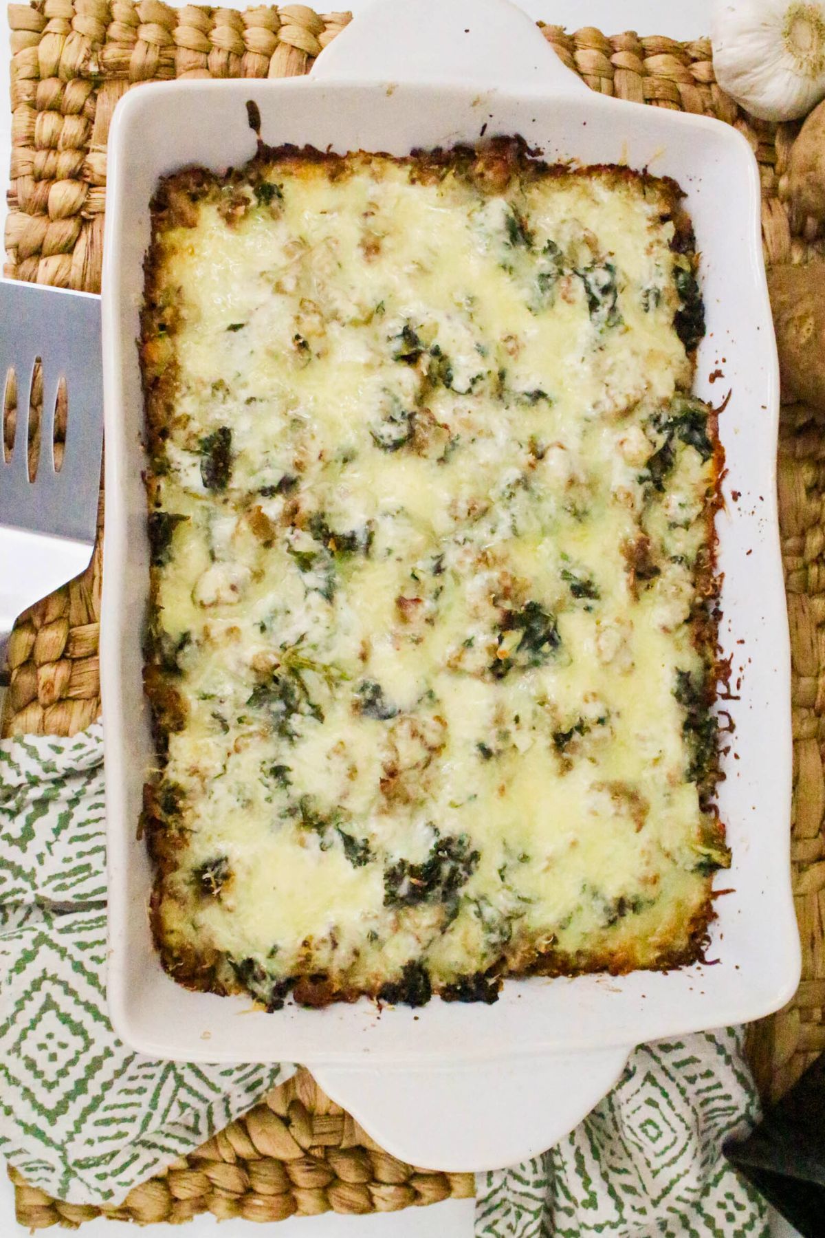 A baked casserole with melted cheese and spinach in a white dish, placed on a woven placemat with a green patterned cloth and a spatula nearby.