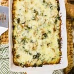 A baked casserole topped with melted cheese and herbs in a rectangular white dish, placed on a woven mat next to a spatula and a patterned napkin.