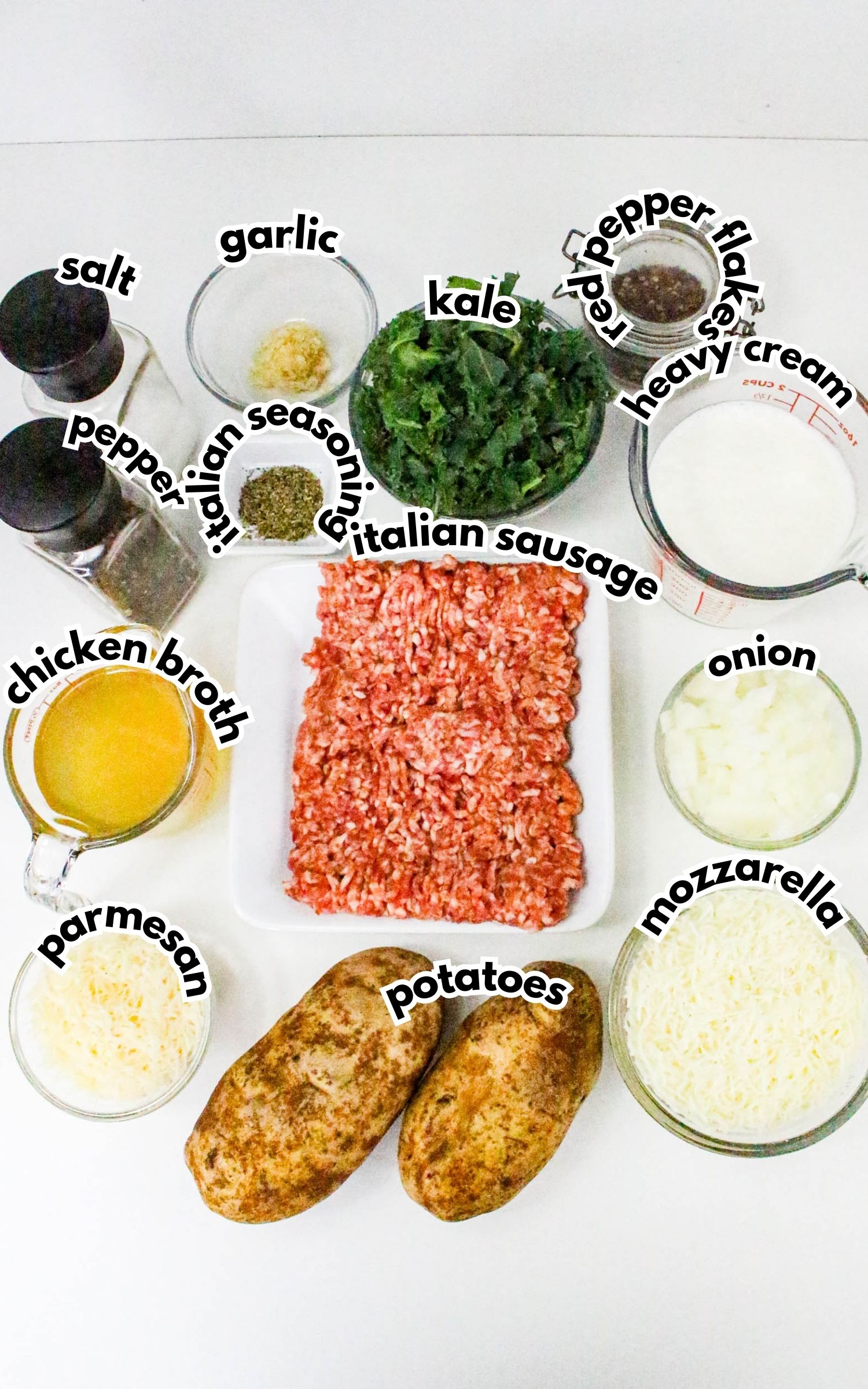 Various labeled ingredients for a recipe are arranged on a white surface, including ground sausage, potatoes, cheeses, seasonings, broth, cream, kale, onion, garlic, salt, and pepper.