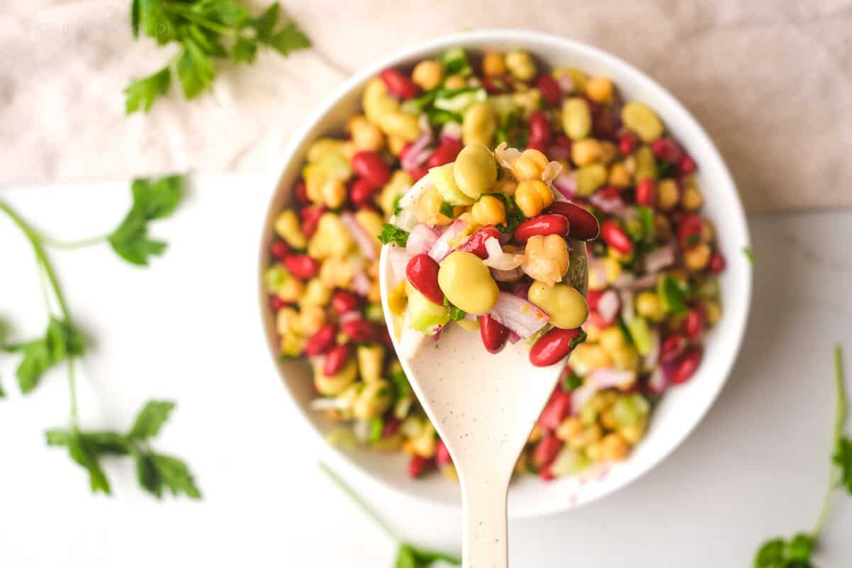 A white spoon holds a mixed bean salad with kidney beans, chickpeas, edamame, red onion, and parsley over a bowl of the same salad.