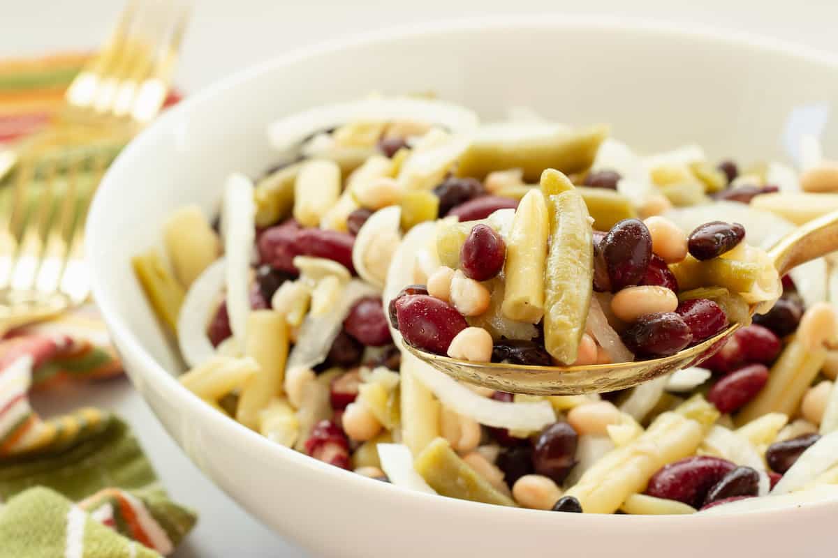 A close-up of a bowl of three bean salad with green beans, kidney beans, black beans, white beans, and sliced onions, served with a spoon.