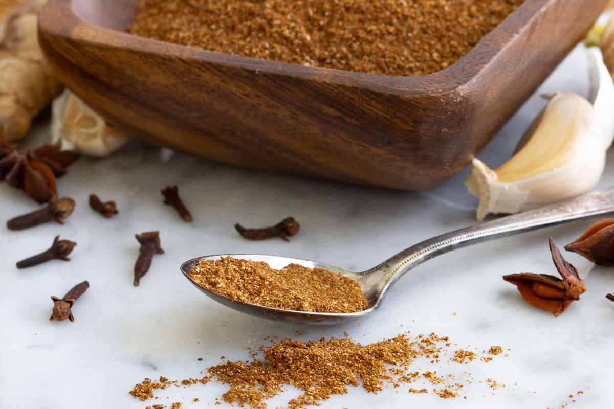 A spoonful of ground spice blend on a marble surface, with a wooden bowl of more spice and scattered cloves, star anise, garlic, and ginger in the background.