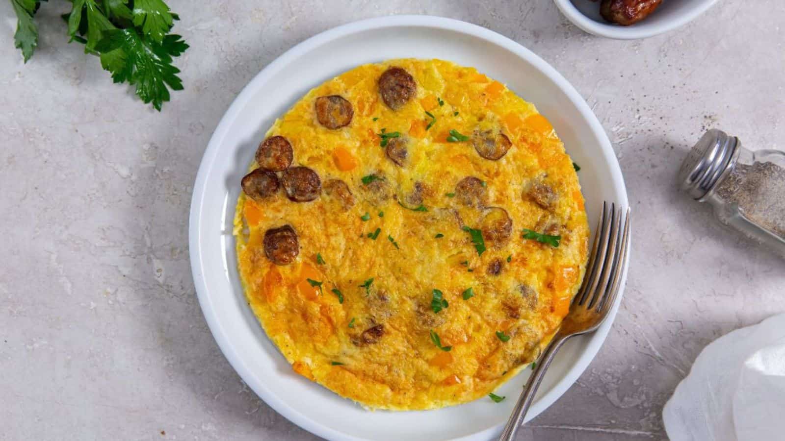 A round omelette with sausage slices and chopped herbs is served on a white plate with a fork, next to a bunch of parsley, a salt shaker, and a small bowl.
