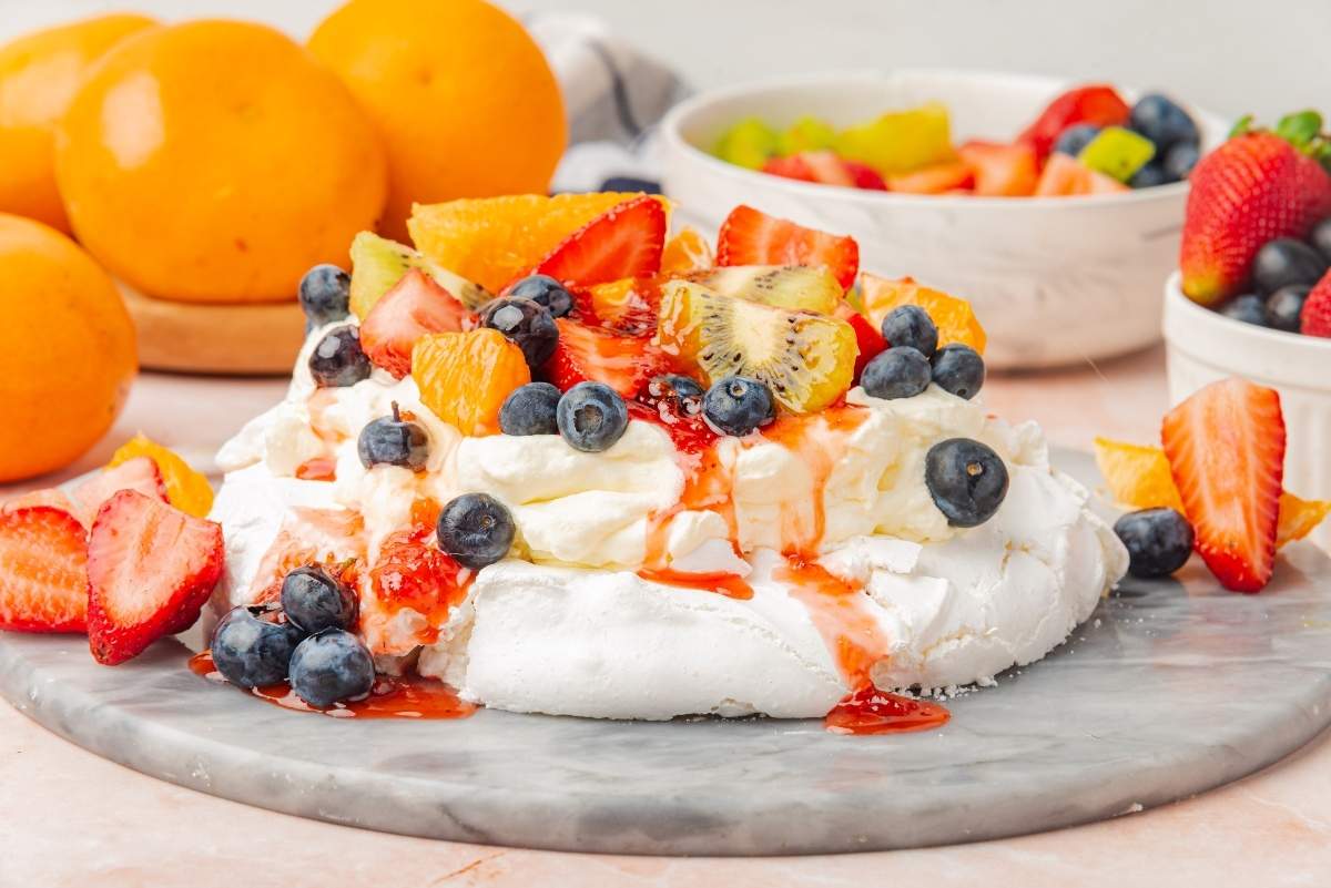A pavlova dessert topped with whipped cream, sliced strawberries, kiwi, blueberries, and orange segments on a marble surface, with bowls of fresh fruit in the background.