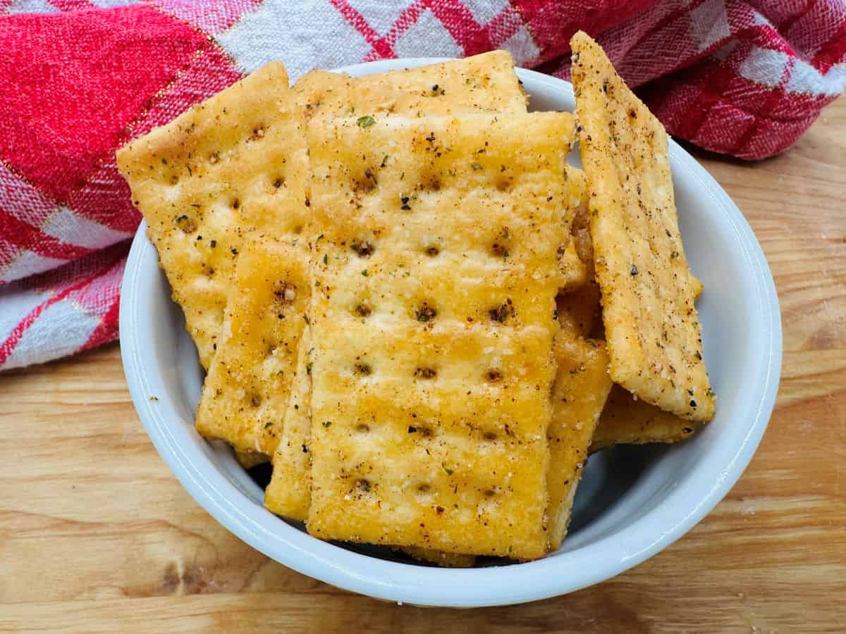 A white bowl filled with seasoned square crackers sits on a wooden surface next to a red and white checkered cloth.