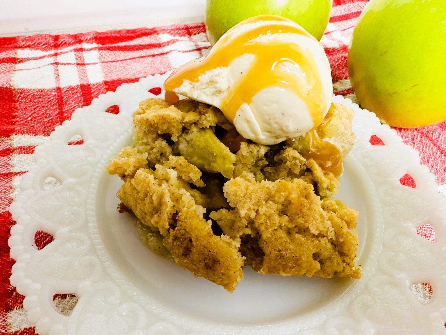 A serving of apple cobbler topped with vanilla ice cream and caramel sauce on a white plate, with two green apples in the background.