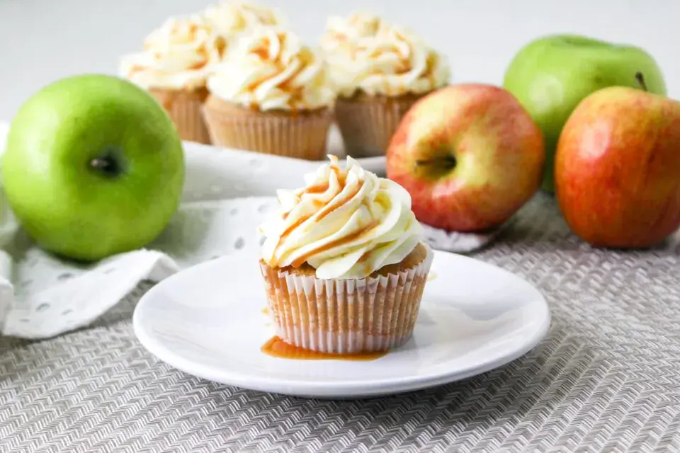 A cupcake with caramel drizzle on a white plate, surrounded by green and red apples, with more cupcakes in the background.