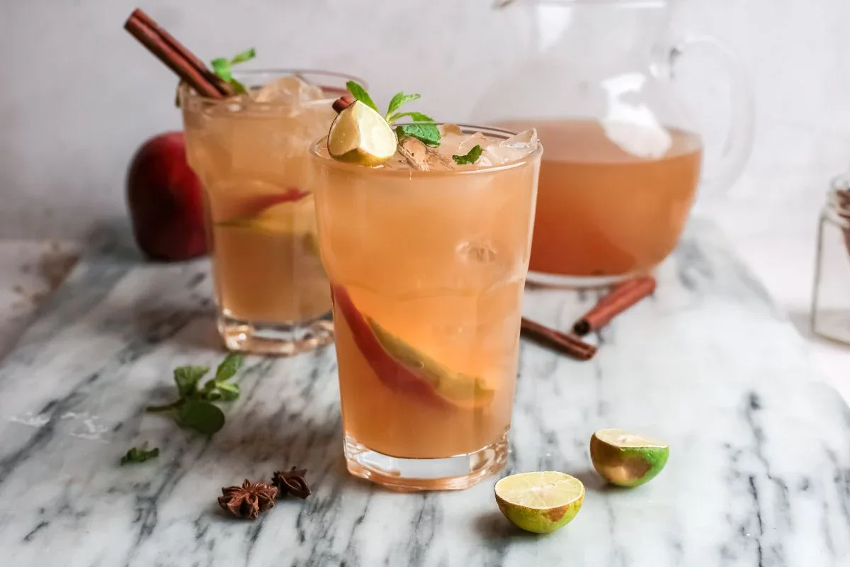 Two glasses of iced apple cider with lime wedges, mint, and cinnamon sticks on a marble surface; a pitcher and extra ingredients are in the background.