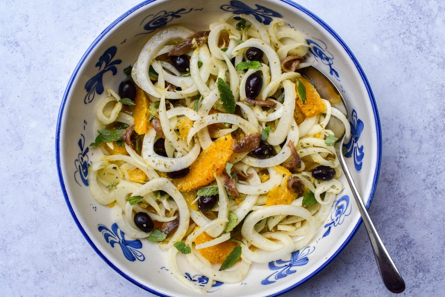 A bowl of salad with sliced fennel, orange segments, black olives, anchovies, fresh herbs, and a fork on the side.