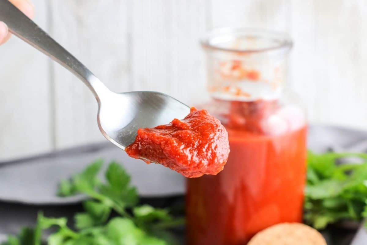 A spoonful of tomato paste held in front of a glass jar filled with more tomato paste, with fresh herbs in the background.
