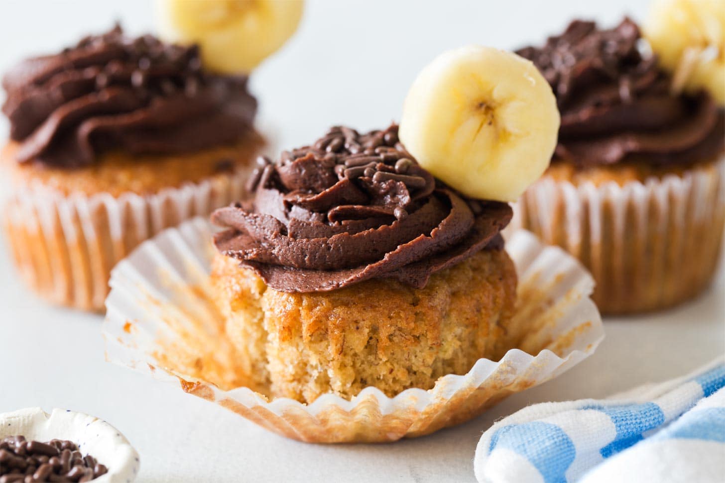 Banana cupcake with chocolate frosting and chocolate sprinkles, topped with a banana slice, in a white cupcake liner. Other similar cupcakes are in the background.