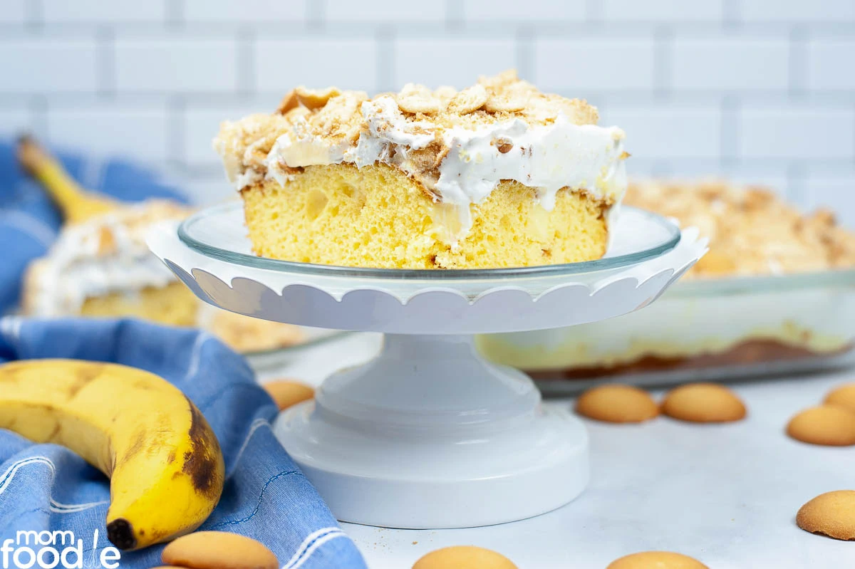 A slice of banana pudding cake with whipped topping and crushed vanilla wafers sits on a glass cake stand, surrounded by bananas and vanilla wafer cookies.