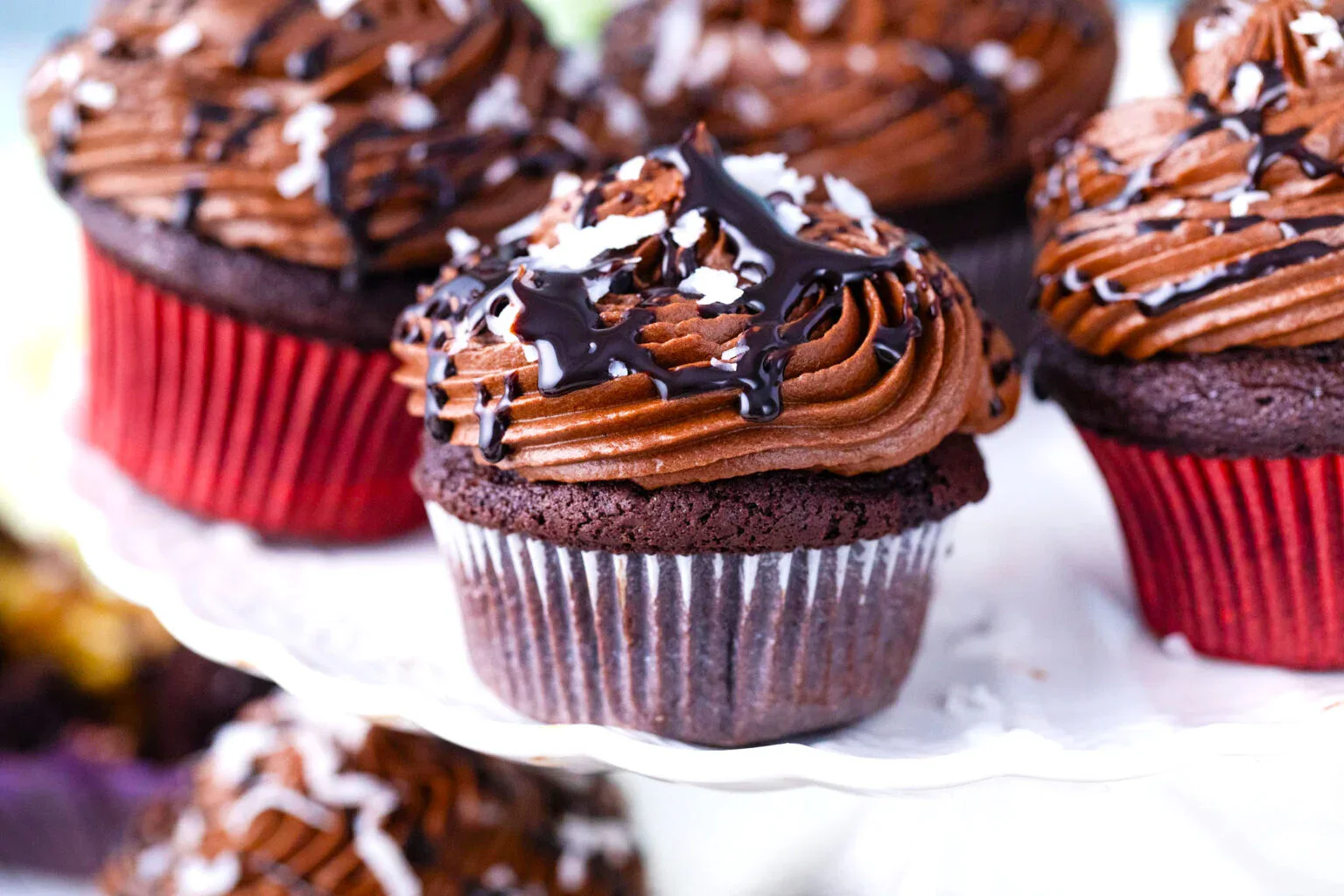 Chocolate cupcakes with swirled chocolate frosting, chocolate drizzle, and shredded coconut on top, displayed on a white cake stand.