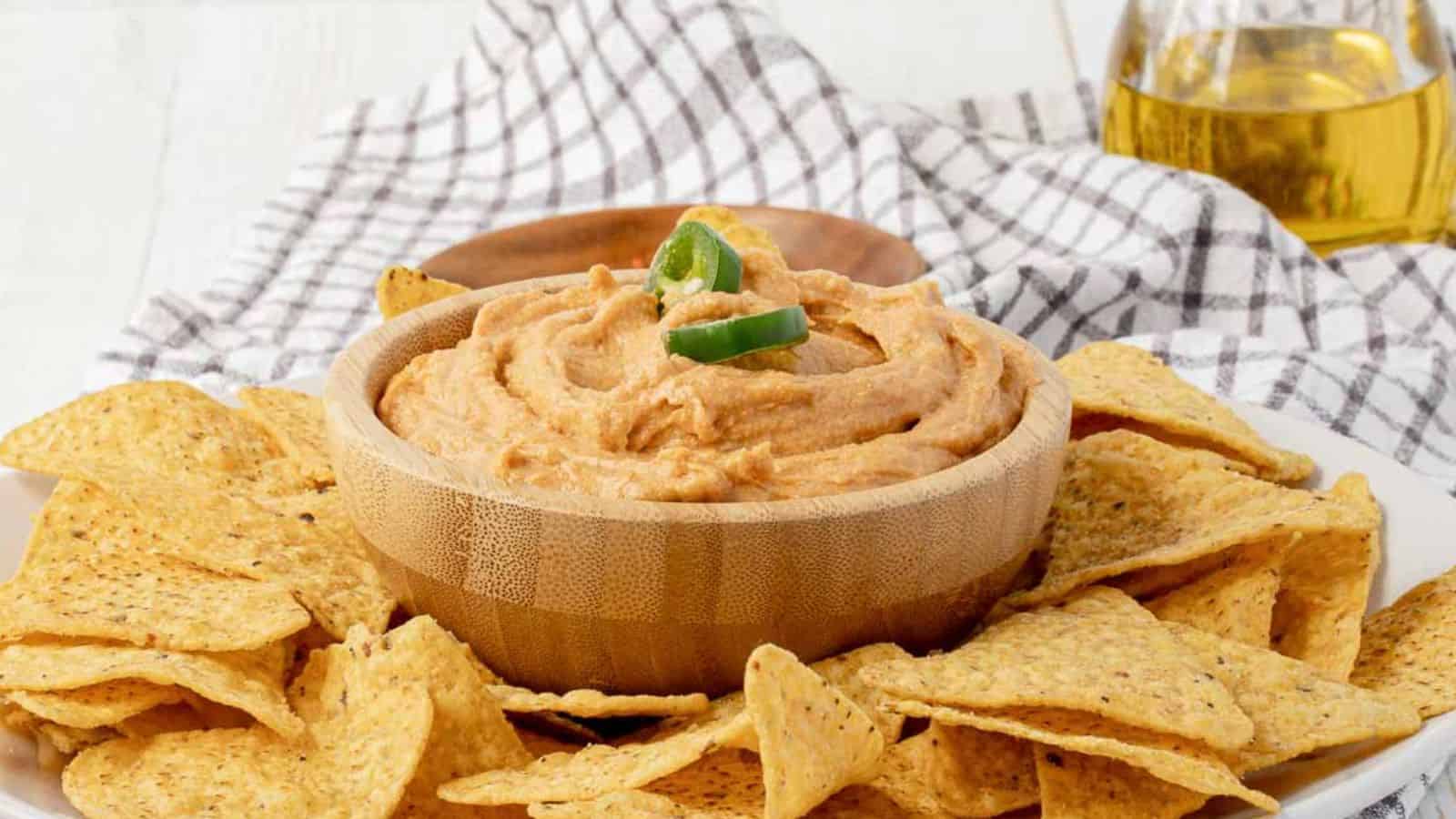 A wooden bowl of creamy dip topped with sliced jalapeños, surrounded by tortilla chips, sits on a white plate with a checkered cloth and a glass of drink in the background.