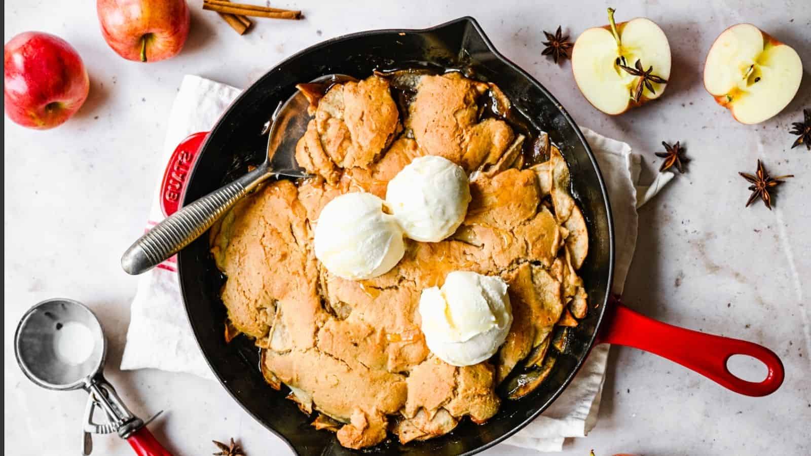 A skillet apple cobbler topped with three scoops of vanilla ice cream, surrounded by fresh apples, spices, and an ice cream scoop on a white surface.