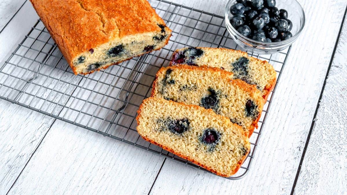 A loaf of blueberry bread sits on a cooling rack with three sliced pieces next to a small glass bowl of fresh blueberries on a white wooden table.