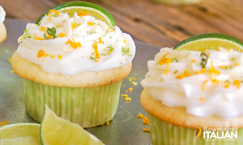 Cupcakes with white frosting, garnished with lime slices and citrus zest, are displayed on a tray.