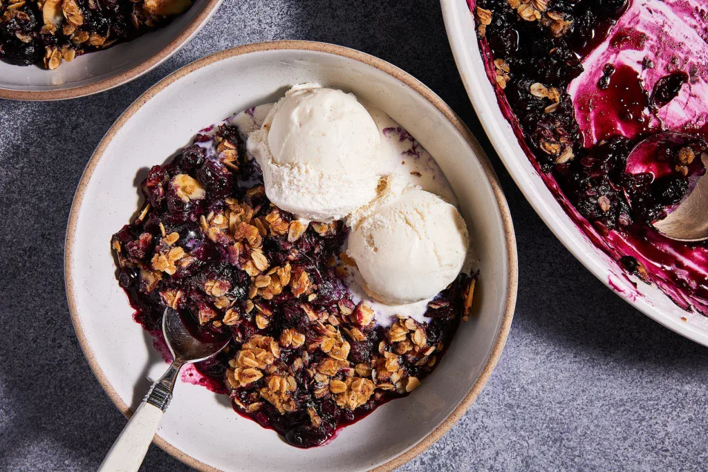 A bowl of blueberry crisp topped with two scoops of vanilla ice cream, with a spoon and a baking dish of crisp nearby.